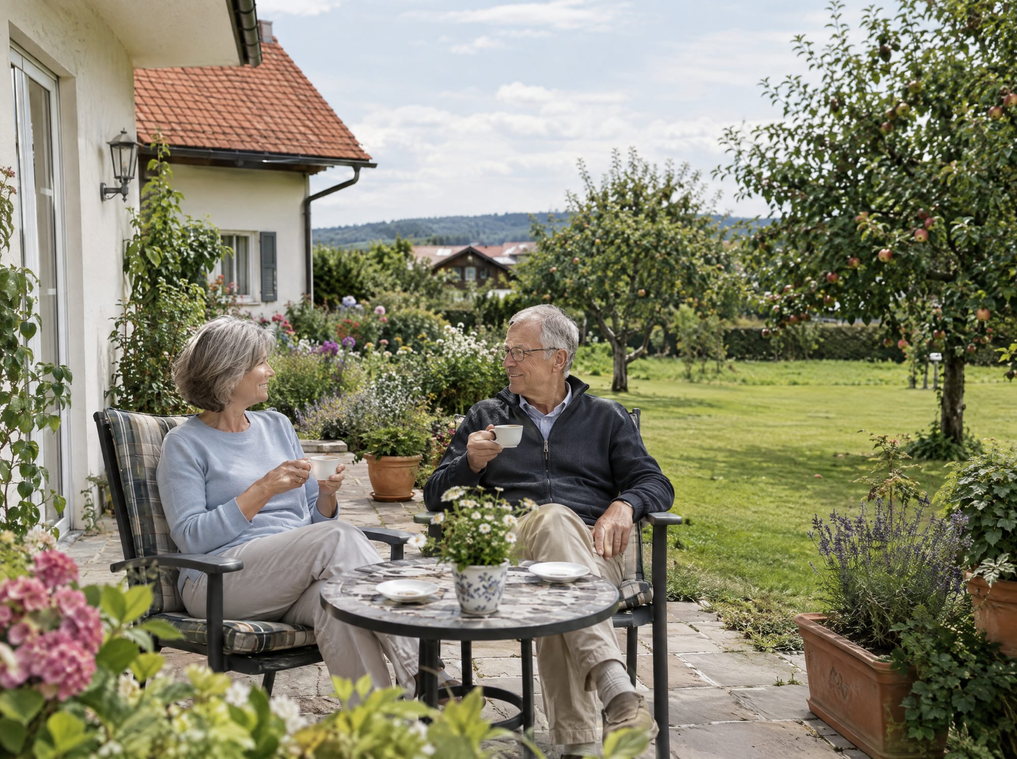 Älteres Ehepaar beim Kaffee auf der Terrasse seines Hauses, dahinter ein großer Garten mit Obstbäumen